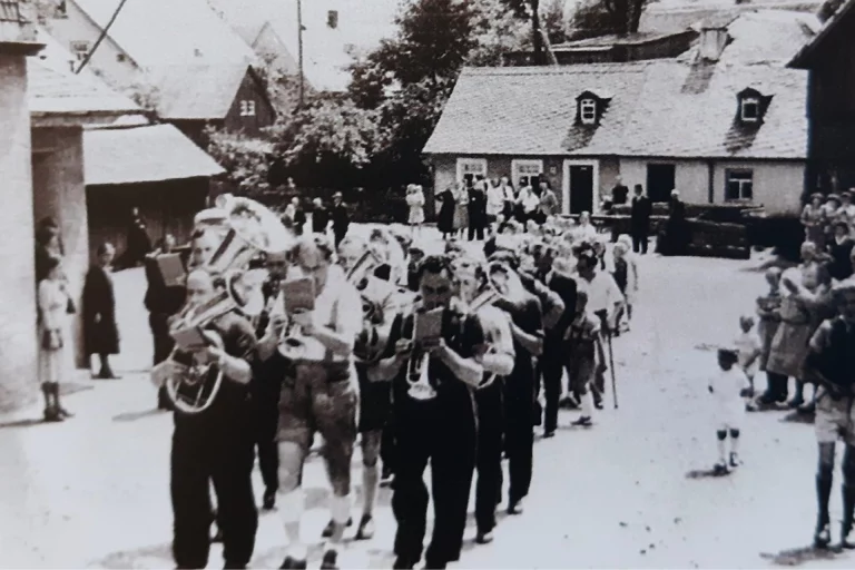Blaskapelle führt historischen Festzug durch ein Dorf an, Anwohner säumen die Straße