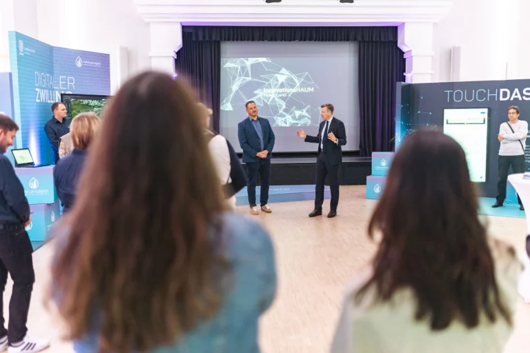 Mann am Rednerpult präsentiert Inhalte auf Leinwand vor Publikum neben Messeständen zum Digitalen Zwilling
