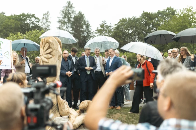 Offizielle und Gäste bei der Auftaktveranstaltung im Freien stehen unter Regenschirmen neben einer Holzskulptur