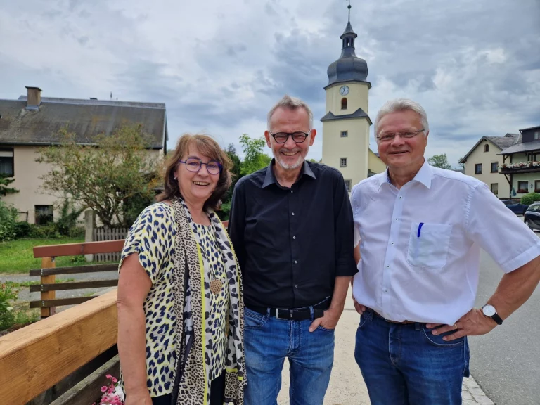 Drei Personen auf einer Brücke vor dem Kirchturm, im Hintergrund Wohnhäuser