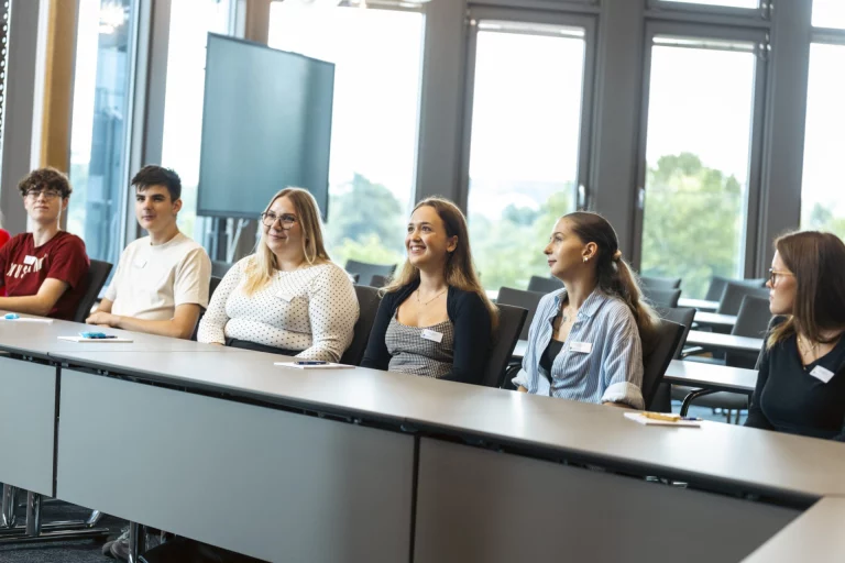 Jugendliche und junge Erwachsene mit Namensschildern sitzen an Tischreihen im Besprechungsraum