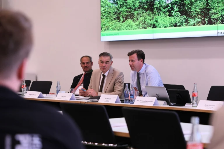 Männer am Podium diskutieren vor Präsentation mit Waldbildern im Sitzungssaal