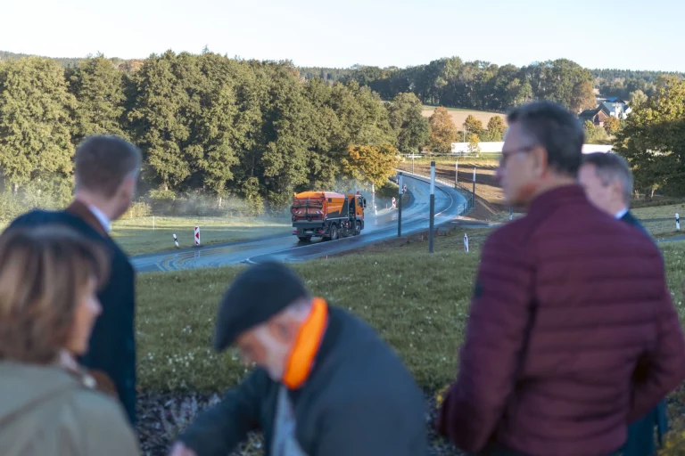 Mehrere Personen beobachten Asphaltarbeiten an einer Straße, im Hintergrund ein Fahrzeug der Straßenbaukolonne