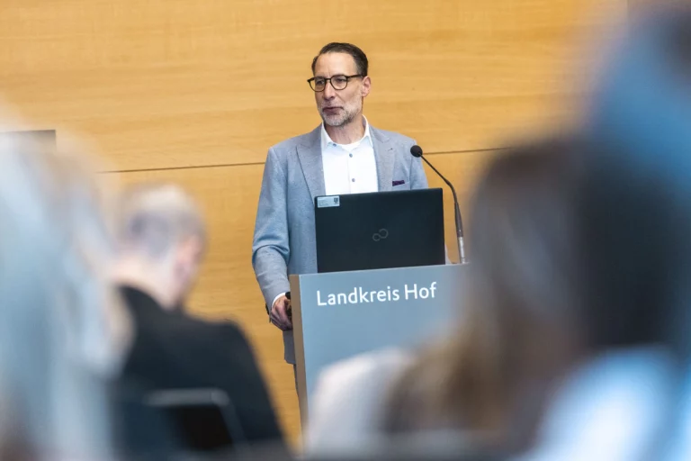 Mann im Anzug spricht am Rednerpult mit Mikrofon vor Publikum, im Hintergrund Schriftzug Landkreis Hof