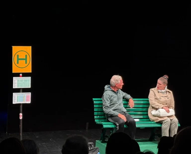 Theaterszene mit zwei Personen im Gespräch auf einer Bank an einer Bushaltestelle