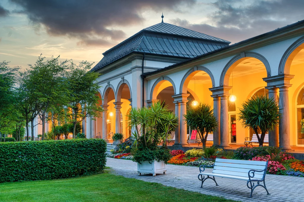 Wandelhalle im Kurpark Bad Steben in der Abenddämmerung, beleuchtete Arkaden und Blumenbeete