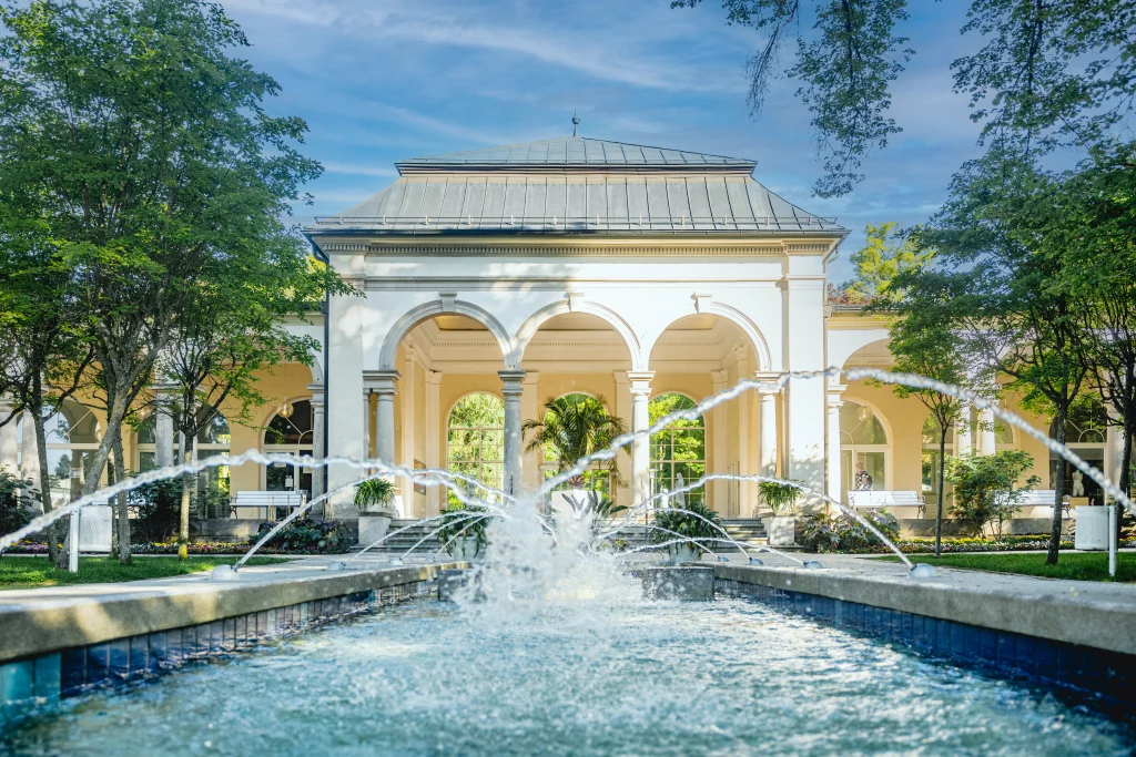Wandelhalle im Kurpark Bad Steben bei Sonne, davor ein Wasserbecken mit Springbrunnen