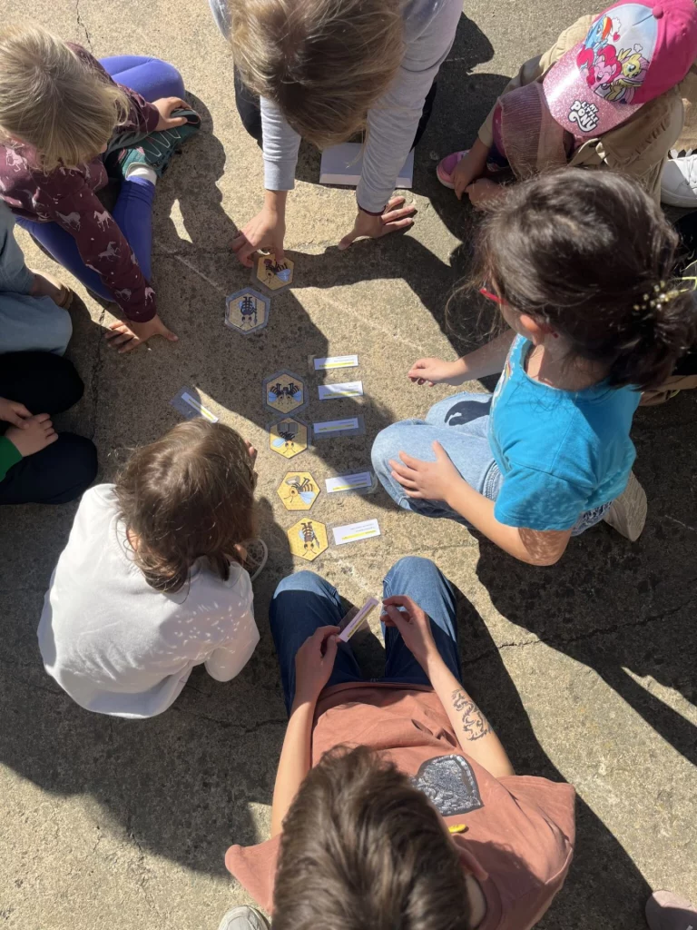 Kinder sitzen im Kreis um sechseckige Spielkarten mit Bienenmotiven auf dem Boden